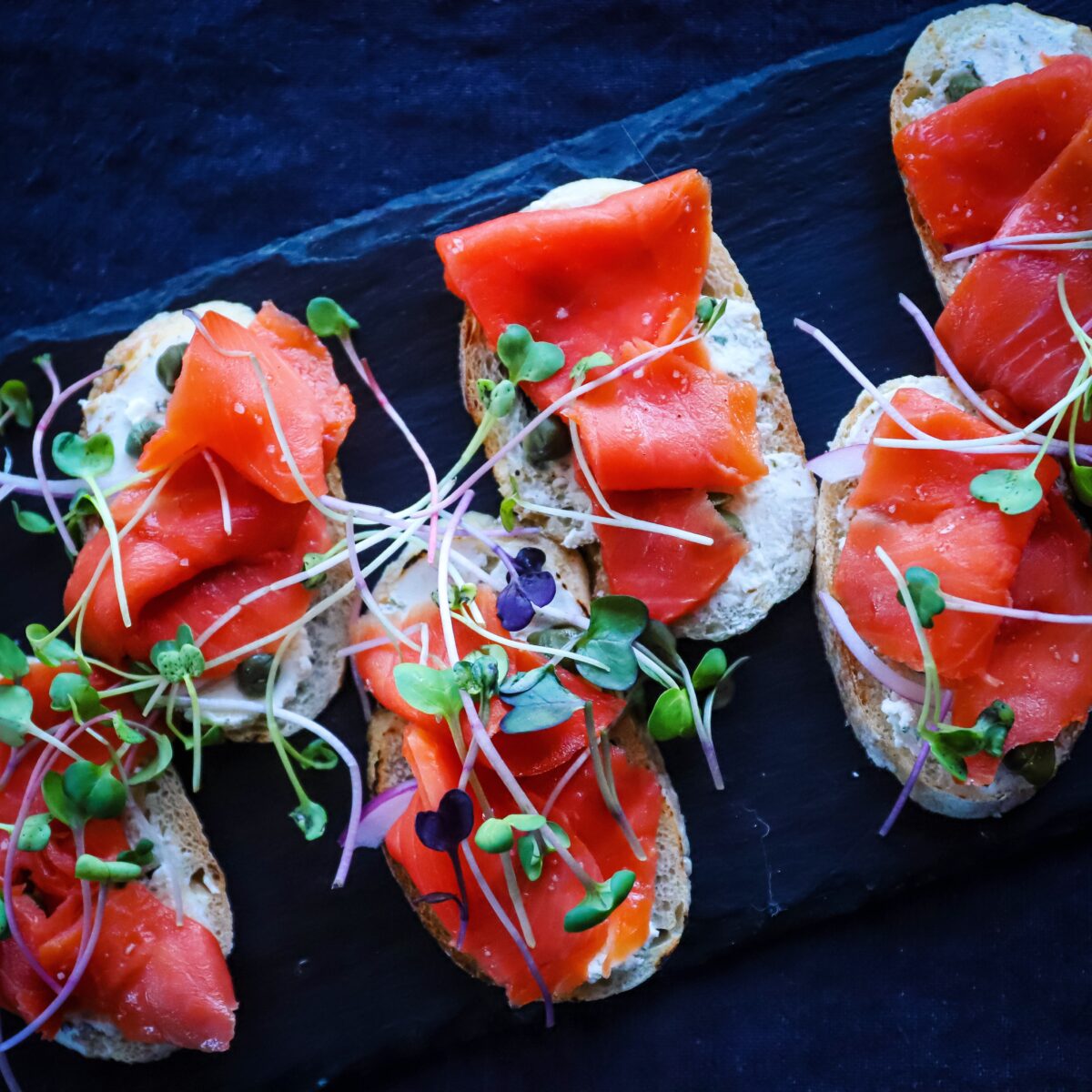Slices of crostini topped with creamy dressing, thin slices of salmon carpaccio and topped with microgreens all on a black tray.