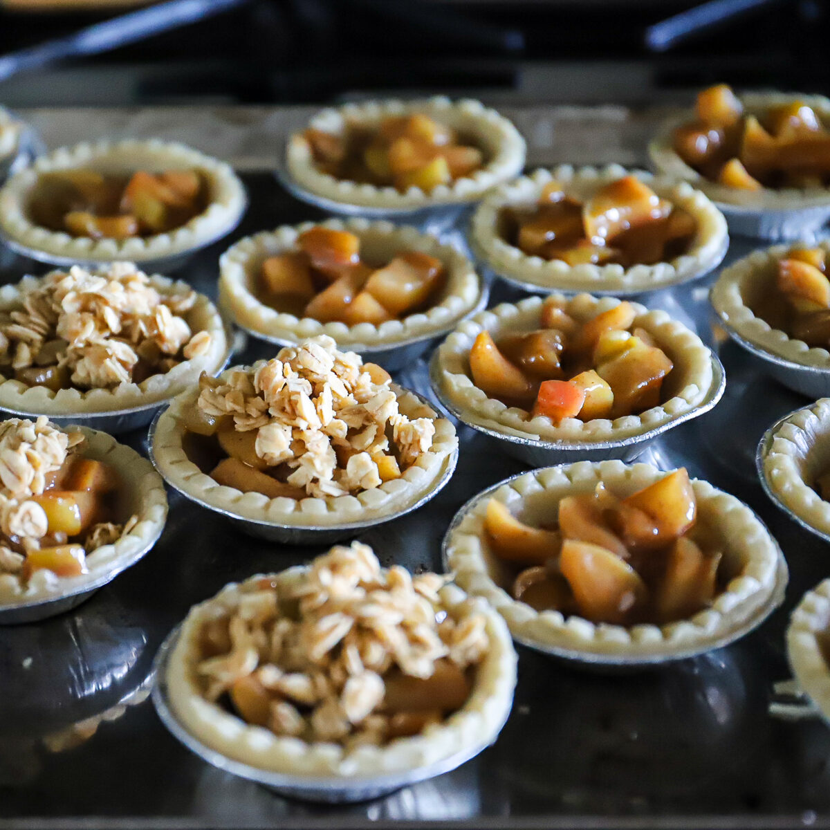 Closeup of raw tartlet shells with apple cinnamon filling in each. Half of them have the crumble topping on top.