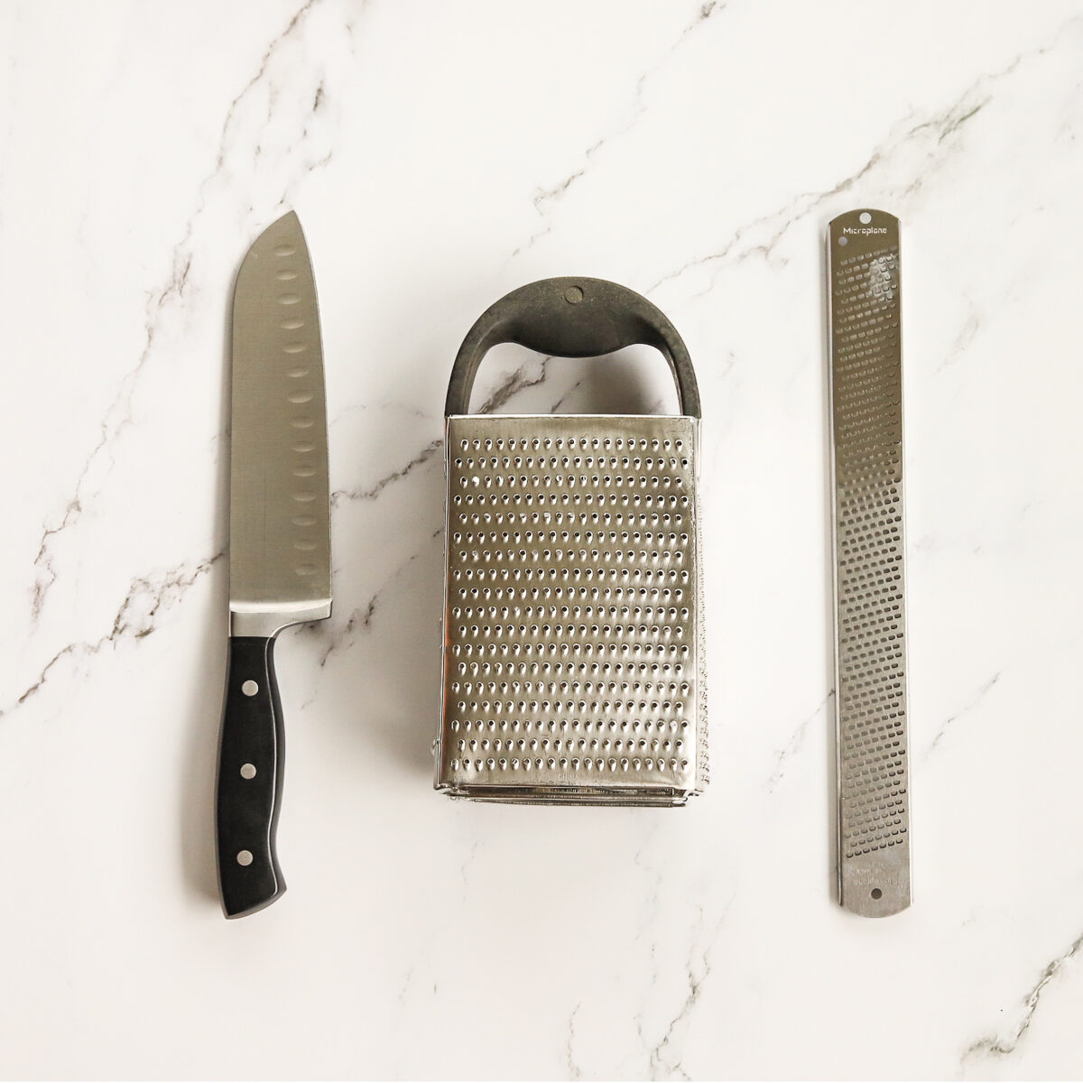 Sharp large chefs knife, box grater and micro plane grater on a white marble countertop.