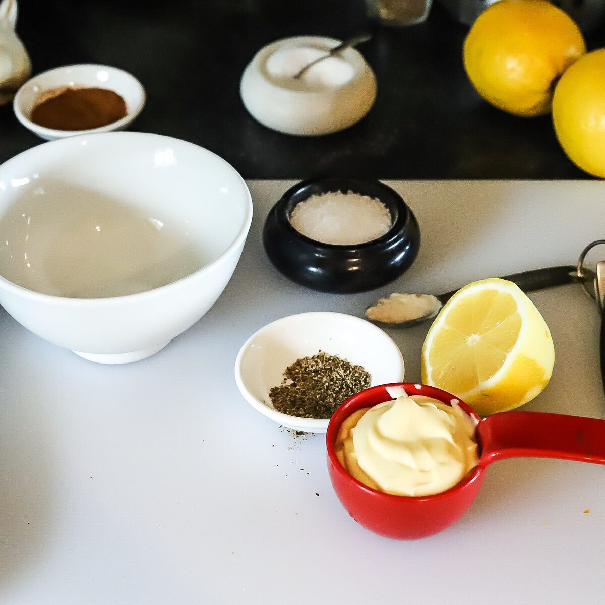 Ingredients for carpaccio dressing measured out in a red cup, and small white and black dishes with an empty bowl and cut lemon halfbeside.