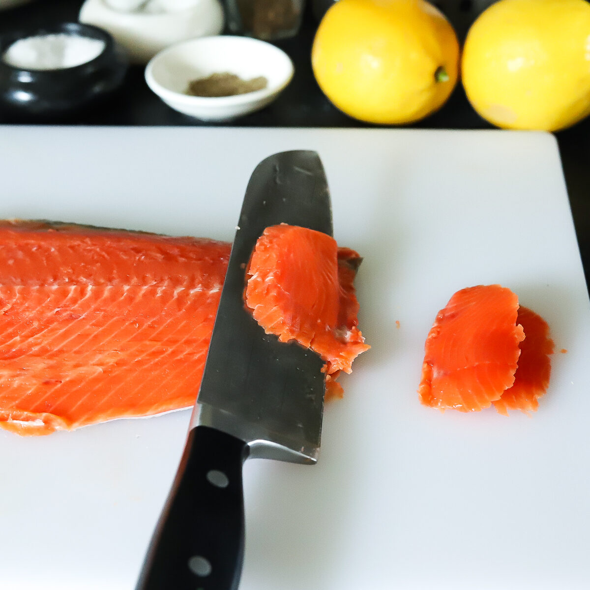 A bright red orange salmon fillet sitting horizontally on a cutting board. There is a large chefs knife slicing the salmon thinly across the grain. 