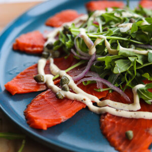 Close up of thinly sliced sockeye salmon drizzled with white mayo based dressing and topped with arugula and a sprinkling of capers and red onions all on a blue plate.