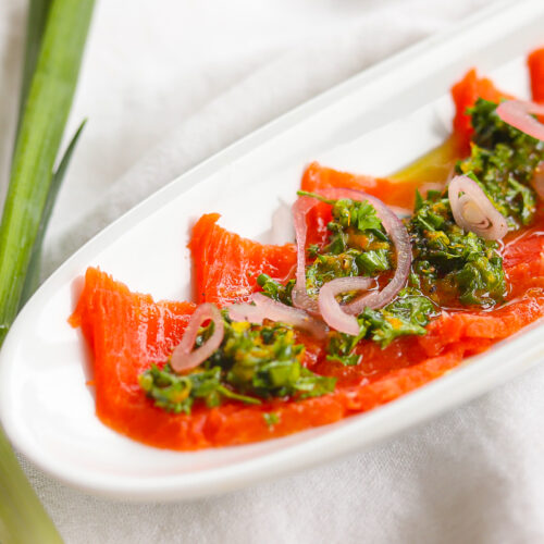 Up close picture of thinly sliced sockeye salmon with chopped herb sauce and pickled shallots on top. It is in a white dish on a white tablecloth with green onions beside the plate.