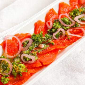 Red thinly sliced salmon on a narrow platter with green herb and citrus dressing down the middle. There are pickled shallots sprinkled over.