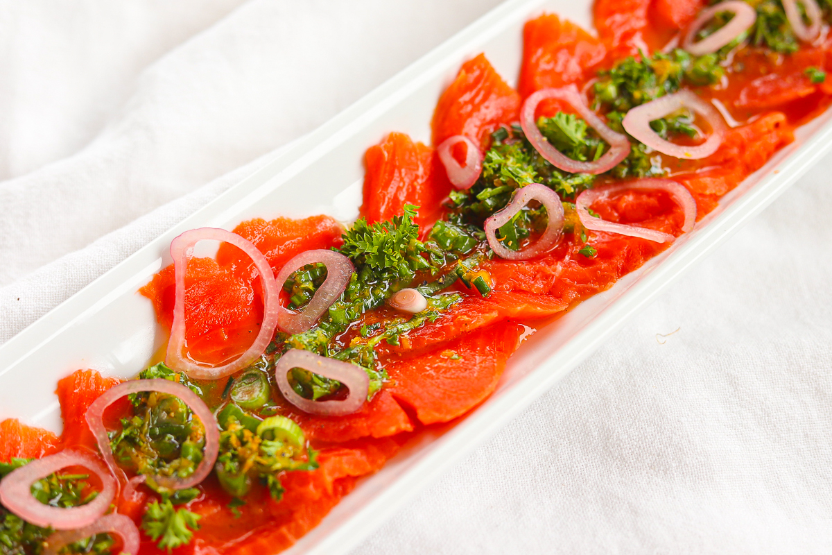 Red thinly sliced salmon on a narrow platter with green herb and citrus dressing down the middle. There are pickled shallots sprinkled over.