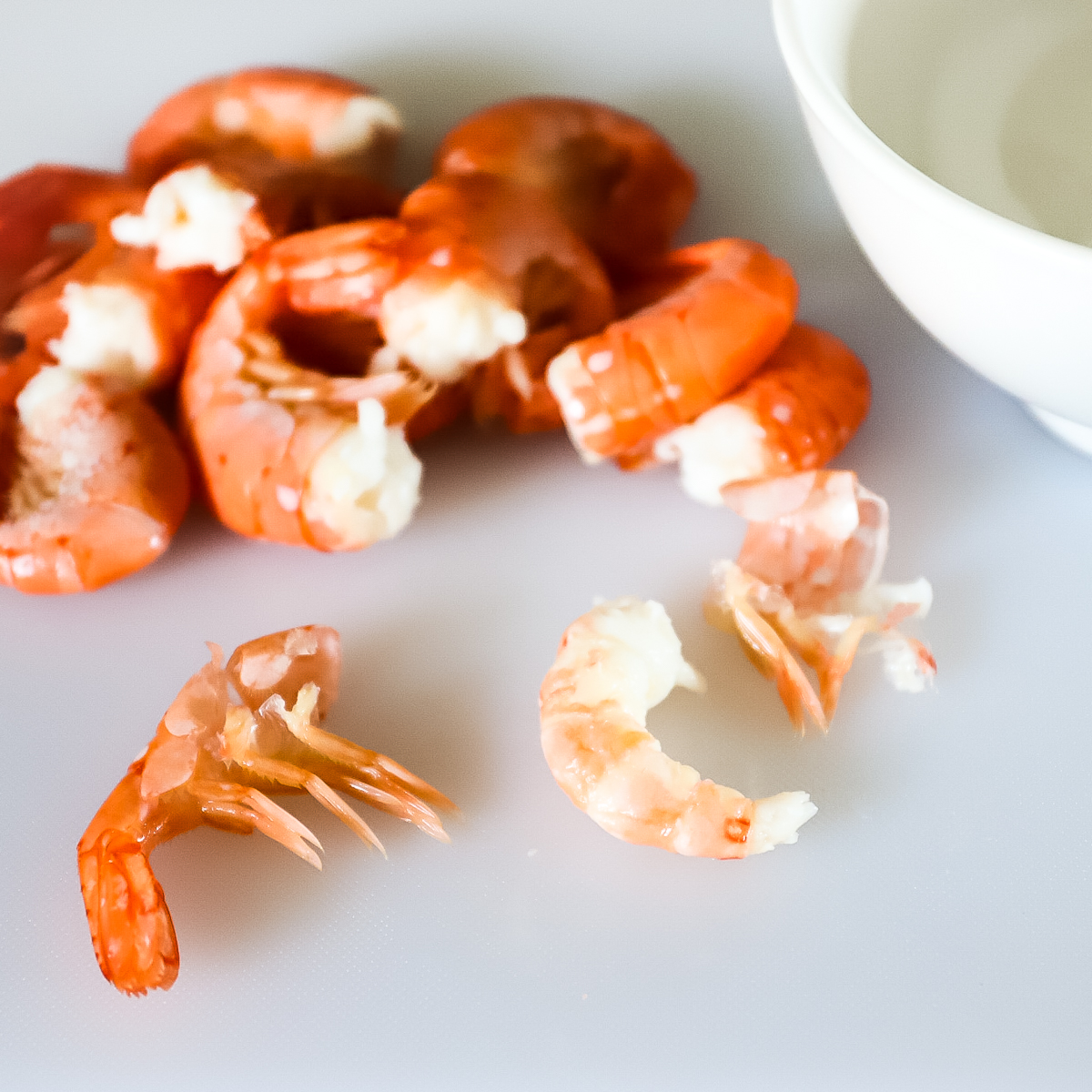 Cooked orange-red prawn tails on a white cutting board with the one closest peeled. You can see the top section of shell peeled off and the bottom half has been pinched and pulled off the bottom of the prawn meat.