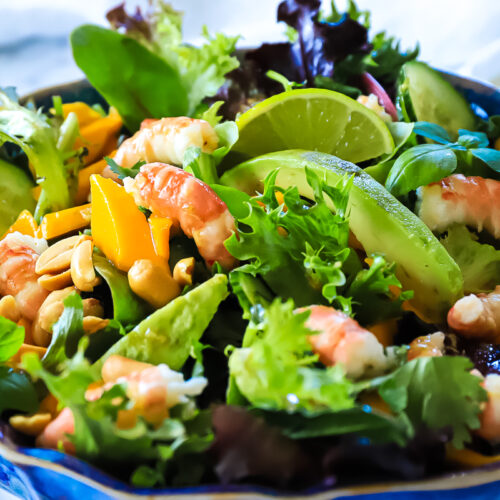 Closeup shot of the finished prawn thai salad showing chunks of mango and avocado, prawns and lime wedges all amongst the mixed greens. It is in a blue bowl.