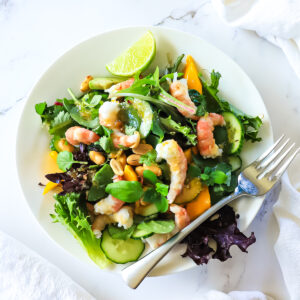 Greens, prawns, mango and lime wedges in a white bowl on a white background with a fork across bowl.