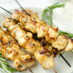 Light colored grilled chicken skewers with rosemary sprigs on each side and a white dip on the upper right hand side.