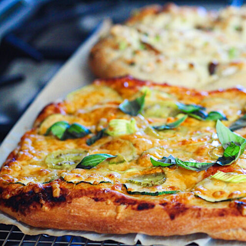 Square pizza's topped with bubbly cheese, basil leaves, zucchini slices and zucchini flowers.