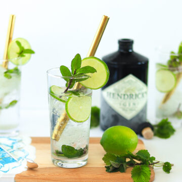 Three tall glasses filled with club soda, mint and lime wheels with gold metal straws in each. Black bottle of hendricks gin in the background and drinks are on a wooden cutting board.