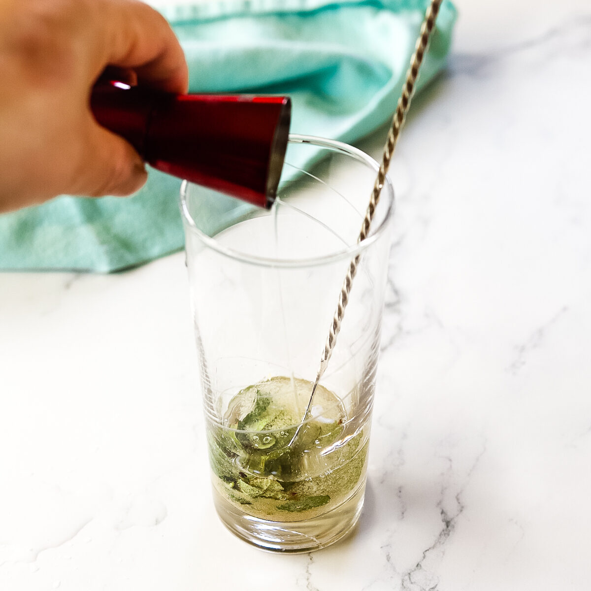 A red shot measure is pouring gin into the muddled mint and sugar in the clear glass.