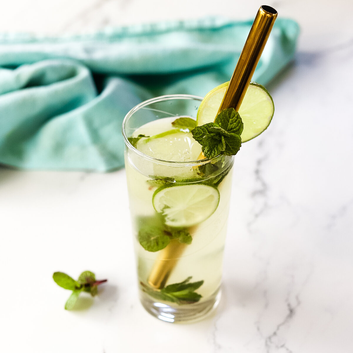 The finished gin mojito on a white countertop with blue tea towel in background. There are pieces of lime and mint floating in the icy cocktail and the metal straw is in diagonally.