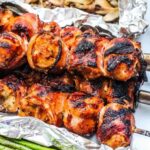 Grilled and charred bacon wrapped chicken cubes on skewers beside green beans on foil.