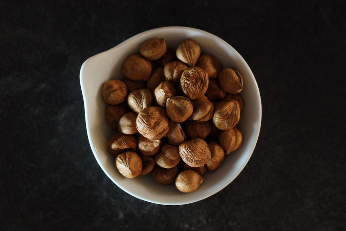 A white bowl of hazelnuts on a black table.
