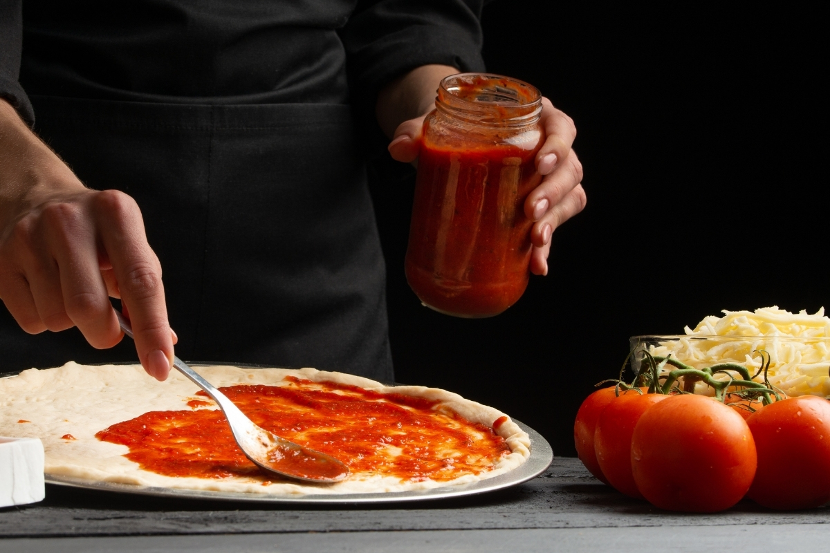 Hand and spoon spreading tomato sauce on the round pizza dough.
