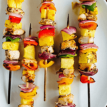 Five grilled kebabs on a white plate. These are Jamaican jerk flavored with pineapple peppers and red onion along with the chicken.
