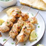 Three light colored cooked chicken kebabs with light green herb dip and lemon pieces around them.