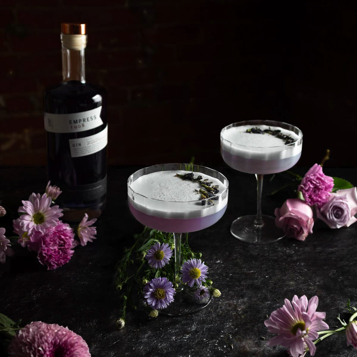 A dark background highlights lavender colored drinks in coupe glasses with a bottle of Empress purple gin and purple flowers all around.
