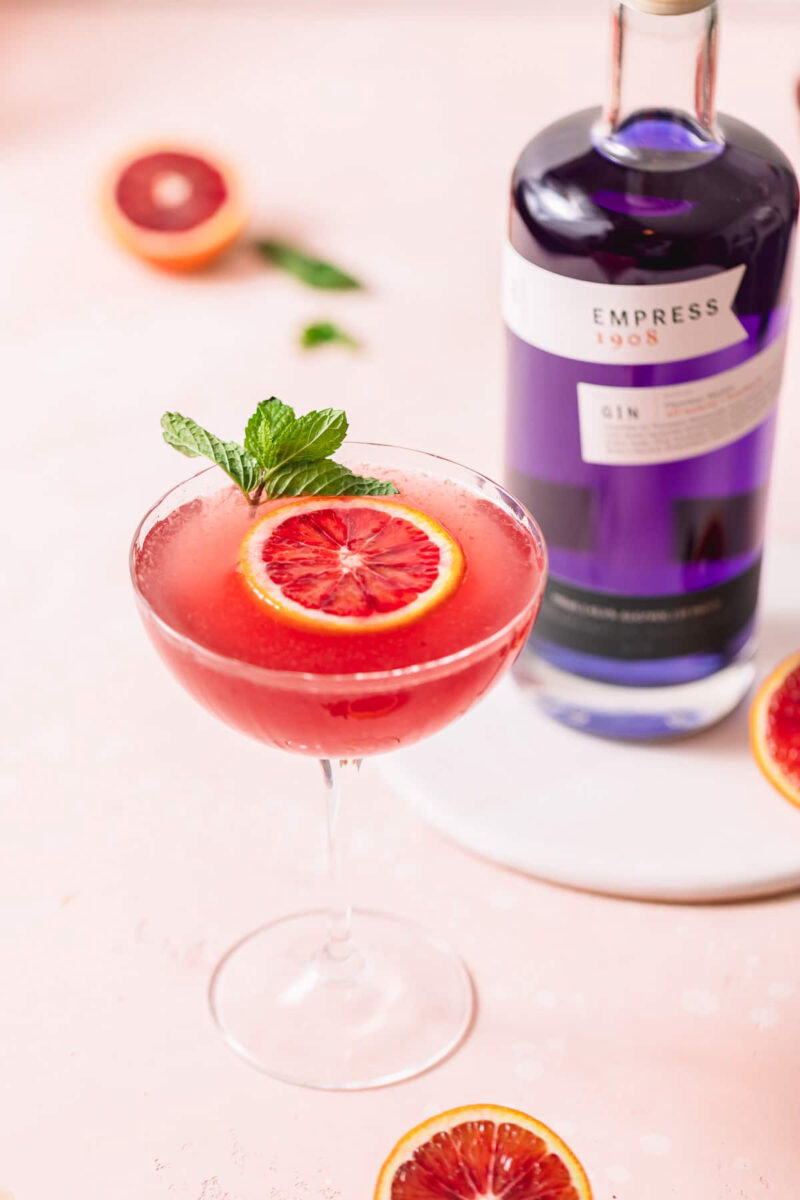 A bright salmon pink drink in a coupe glass with a slice of blood orange floating in it and a mint sprig in the back. A bottle of purple Empress gin in standing to the back right of the glass.