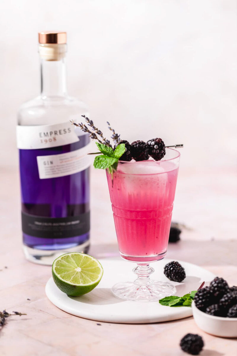 Bottle of purple gin beside a bright pink drink garnished with a pick of blackberries and mint. There is a lime half on the table beside along with more blackberries.