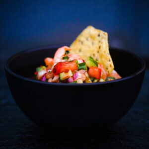 Diced shrimp (camaron), red peppers, cucumber and red onion in a black bowl with a tostada shell stuck in the back of the bowl to scoop with. Background is dark blue black.
