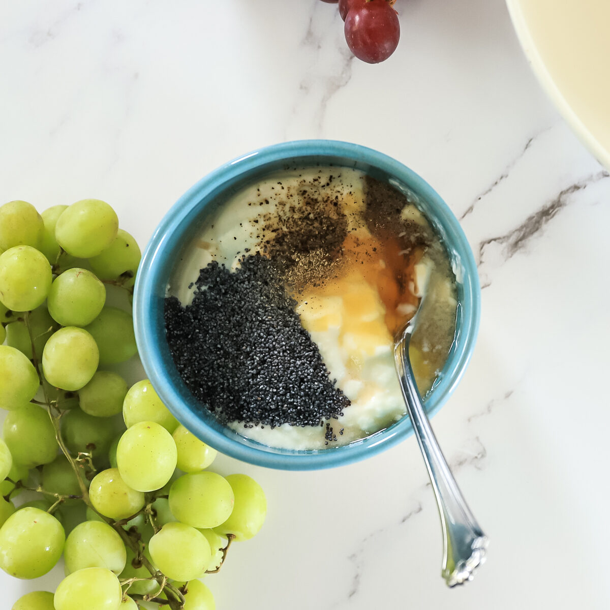 Blue bowl beside green grapes with white yogurt, yellow honey and dark grey poppy seeds in it with a spoon.