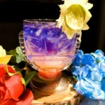 A glass tiki style goblet holds a drink that is purple on top fading to yellow, then orange and last red on the bottom. It is garnished with a mushed lime made to look like the sun.