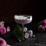 A coupe glass with a pale purple colored frothy gin sour in it. There are pink and purple flowers all around and a dark background.