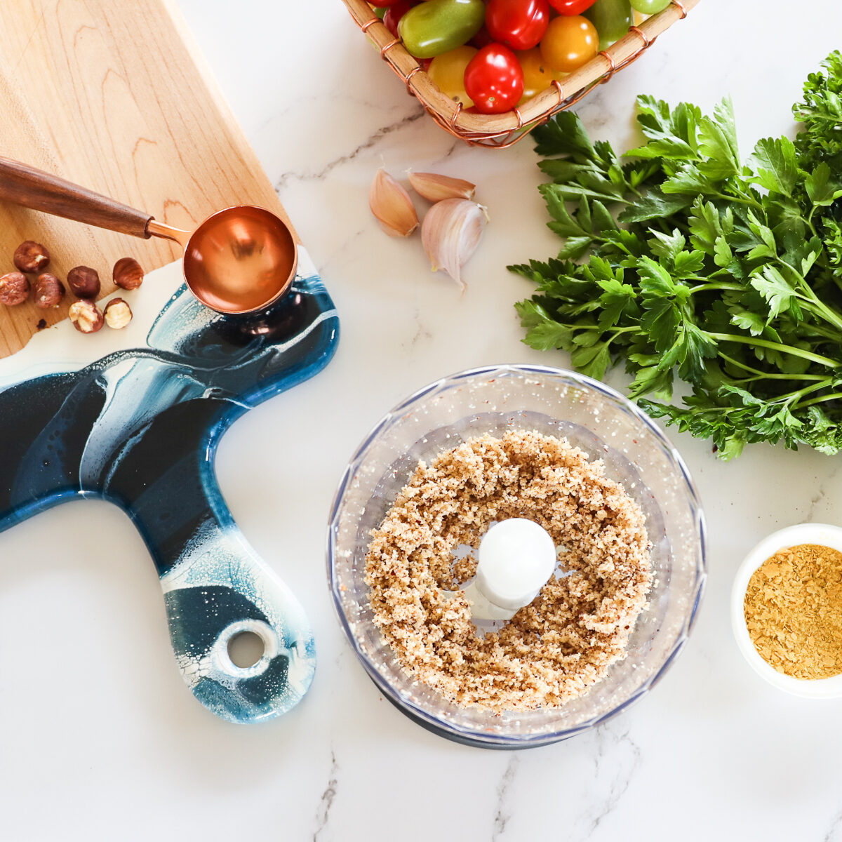 Bowl of food processor with finely ground hazelnuts in it. There are cherry tomatoes, parsley, nutritional yeast, hazelnuts and garlic cloves around it on the white countertop.