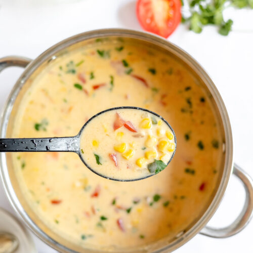Finished Panera corn chowder is pale apricot colored creamy soup with flecks of red, yellow and green in it. There is a spoon full held above the pot to show the thick and creamy texture.