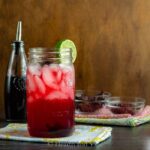 Red hibiscus margarita in a tall mason jar with lots of ice cubes in it and a lime wedge on the top edge of the glass.