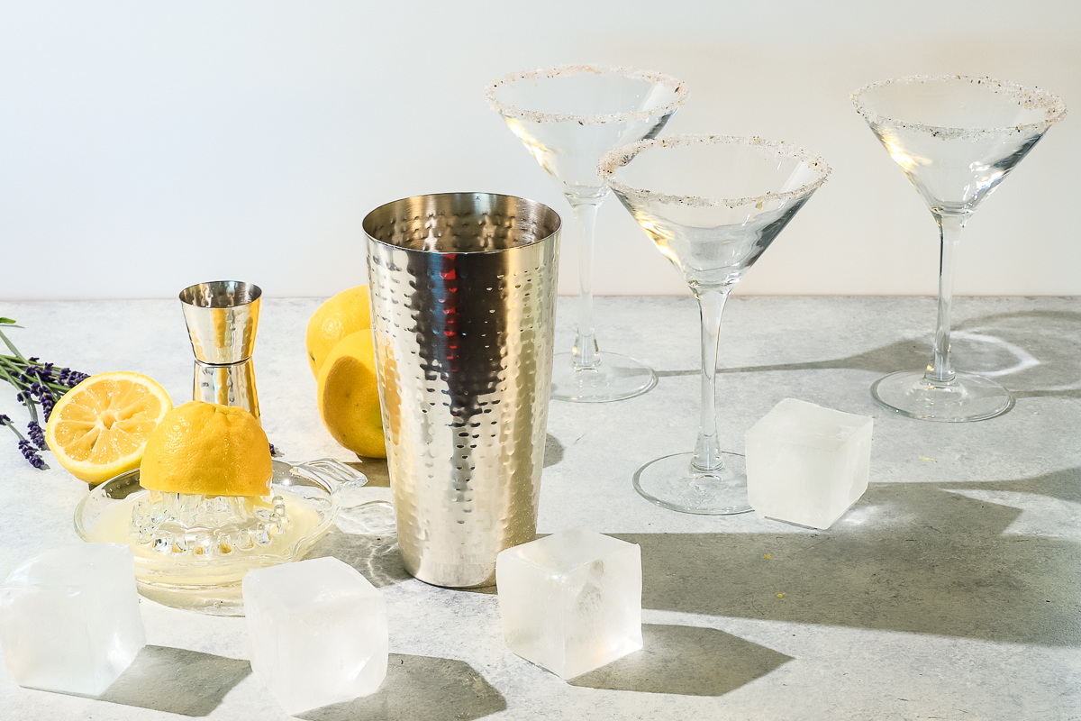 A lemon being juiced on a glass citrus press beside silver cocktail shaker, empty sugared rim martini glasses and scattered ice cubes.