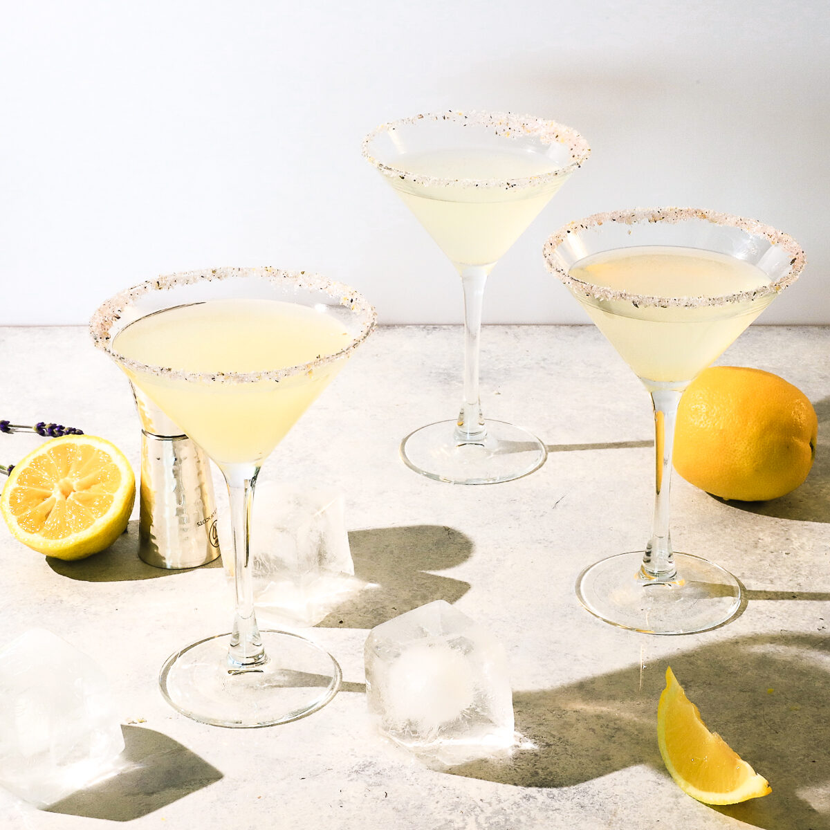 Three martini glasses with lavender sugar rims and pale yellow cocktails poured in them. There is a lemon and cut lemons and ice cubs around on the pale countertop.