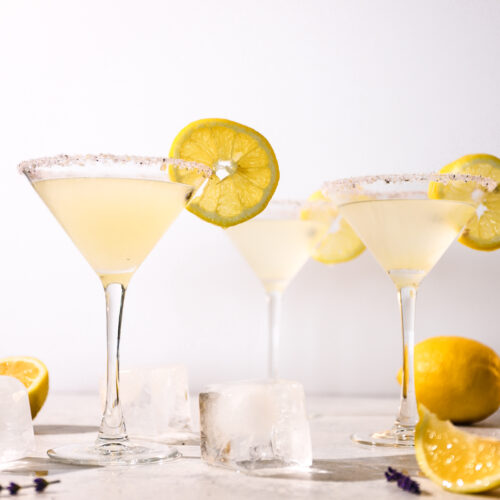 Three pale yellow lavender drinks in martini glasses with a lemon wheel on each.