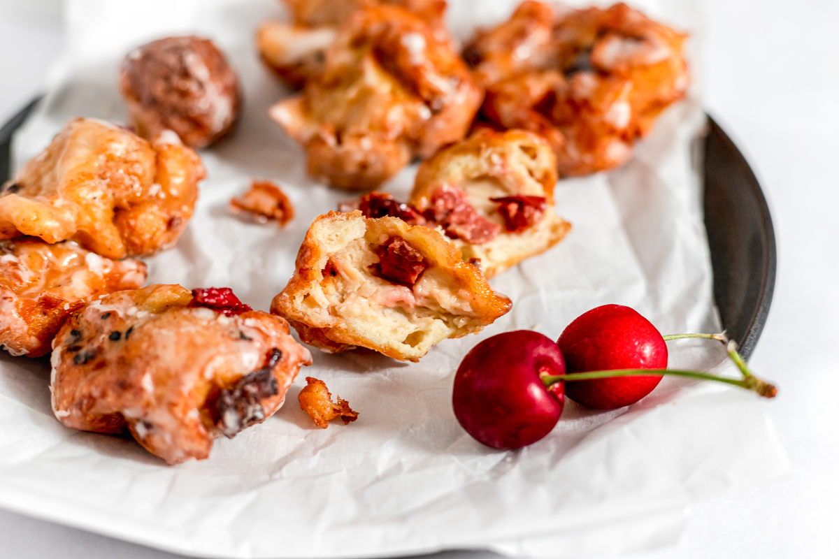 Cherry fritters have a thin icing sugar glaze over them and they are on a parchment lined plate garnished with a few fresh cherries.