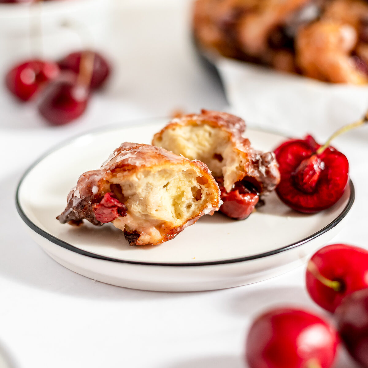 White plate with black rim holds a fritter that is pulled apart to reveal the tender inside texture. Cherries are scattered in front and behind the plate with more fritters in the background.