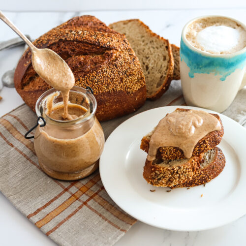 A spoonful of hazelnut puree being spooned out of a glass jar. There is a loaf of bread behind it, a cup of coffee in the top right corner and a white plate of toast topped with the nut puree beside it.