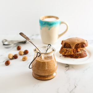 Hazelnut butter in a small glass jar with a spoon in it. There is a blue and white cup of coffee behind and a plate of toast with the nut butter on it.