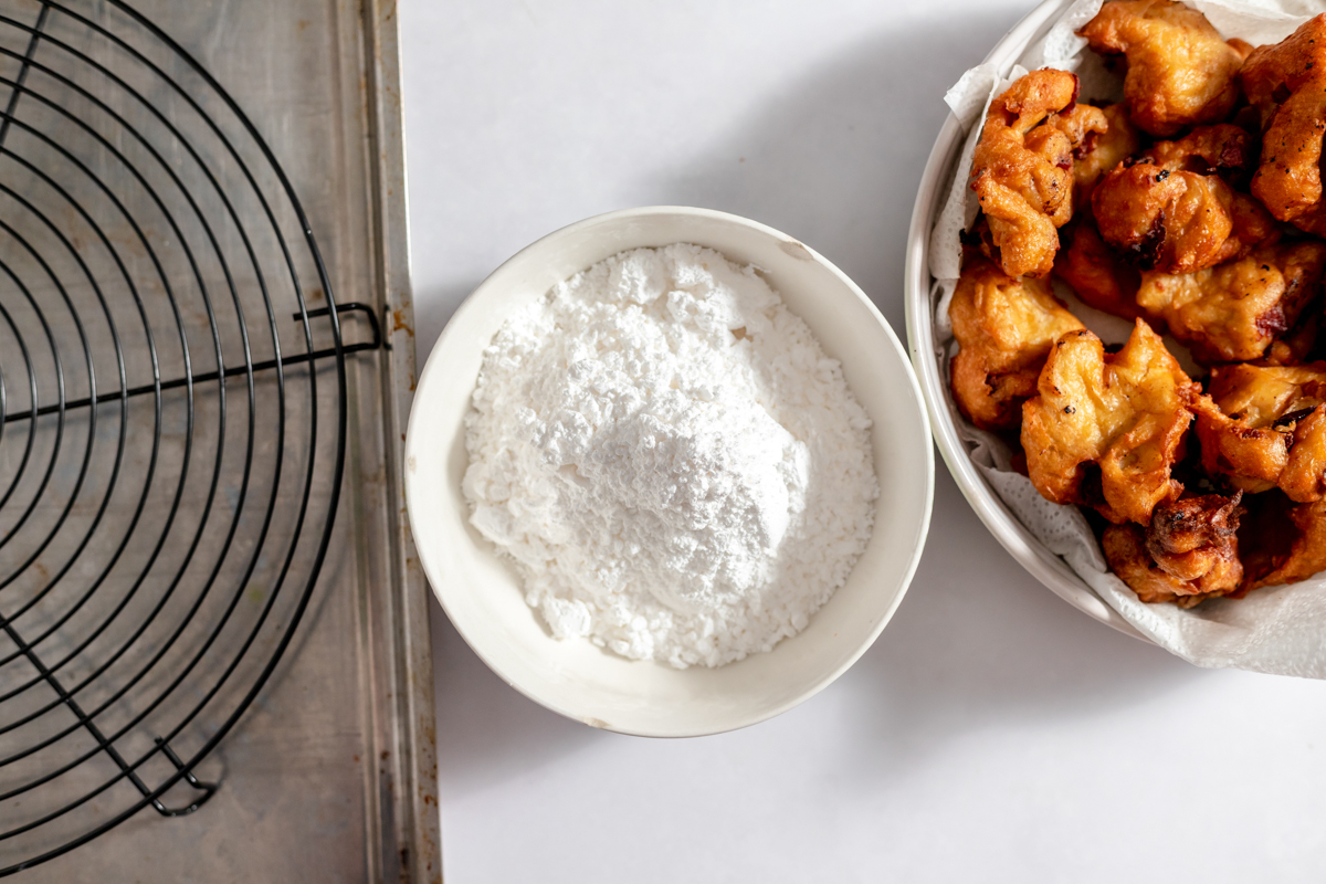 A paper towel lined bowl holds the finished fritters and a smaller bowl of powdered sugar sits beside it.