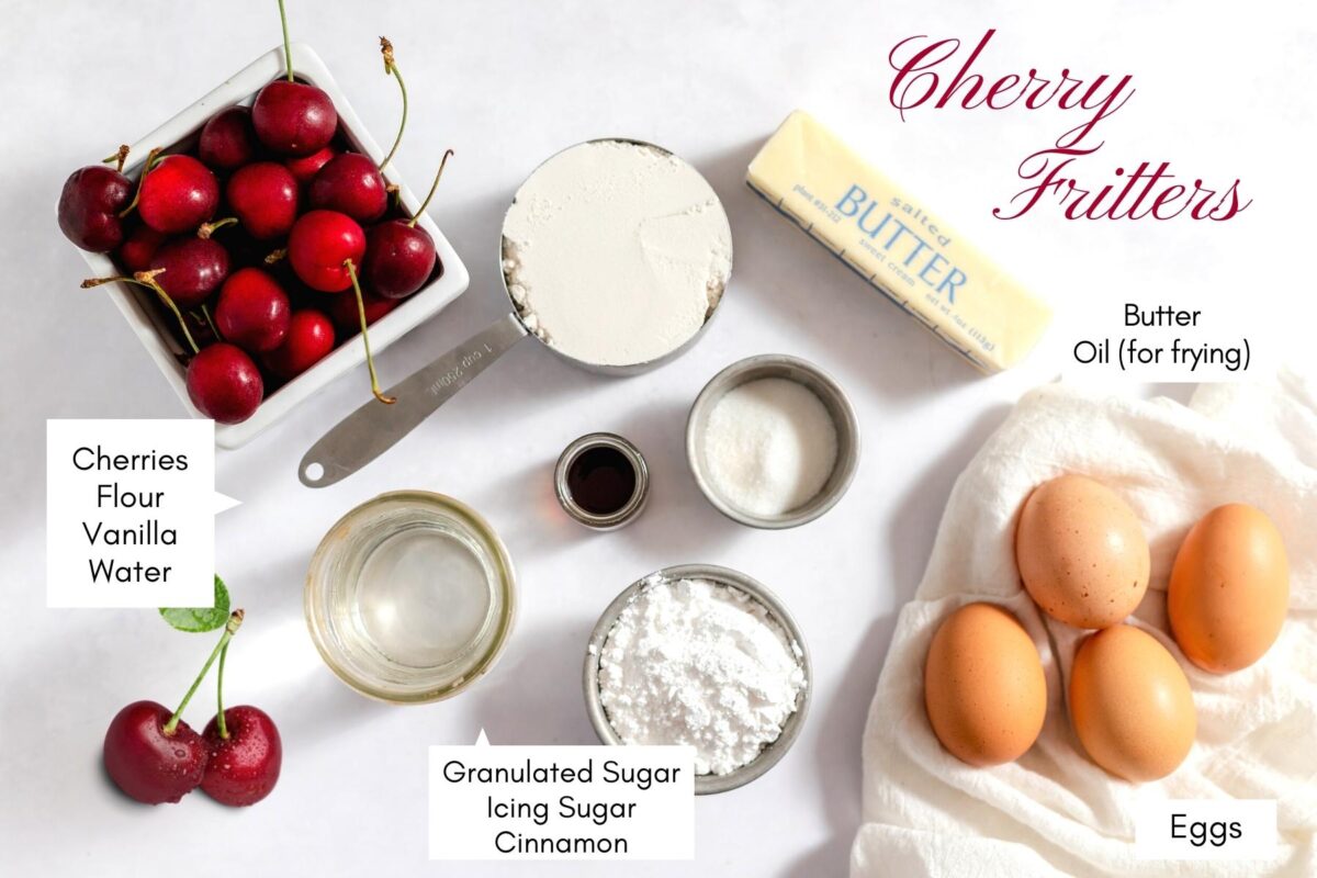 Overhead picture of all the ingredients layed out on a white table with text overlay labelling them. Ingredients include cherries, flour, sugar, butter, eggs, vanilla and powdered sugar.