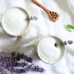 2 mugs of foamy white lavender milk tea on a white tablecloth. There is a wooden honey spoon, a few mint leaves and a small bunch of fresh lavender are laying beside.