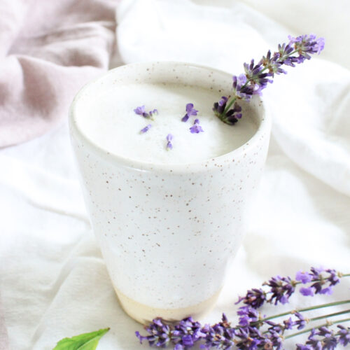 A tall cream coloured mug with frothy lavender tea garnished with lavender buds and a spring of lavender.