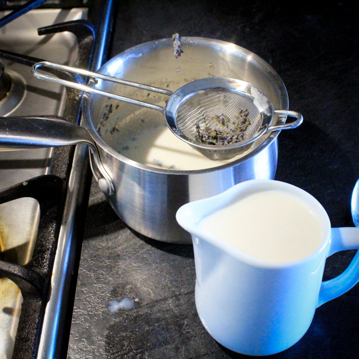 Strained milk tea is in a small white ceramic pitcher. The remaining lavender is in the tea strainer over the saucepan.