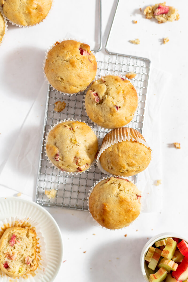Baked muffins on a wire rack with some cut rhubarb in the corner.