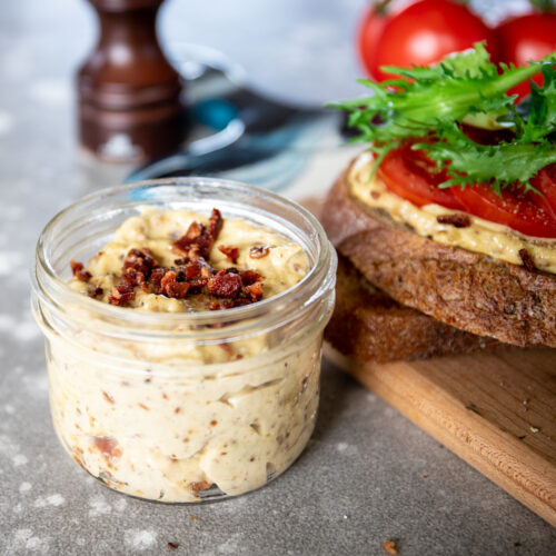 A half pint mason jar holds light tan colored bacon aioli and is topped with crumble bacon. There is a wooden board beside with toast topped with the aioli, tomato slices and lettuce beside. They are all sitting on a mottled grey countertop with a pepper grinder and whole tomatoes in the background.