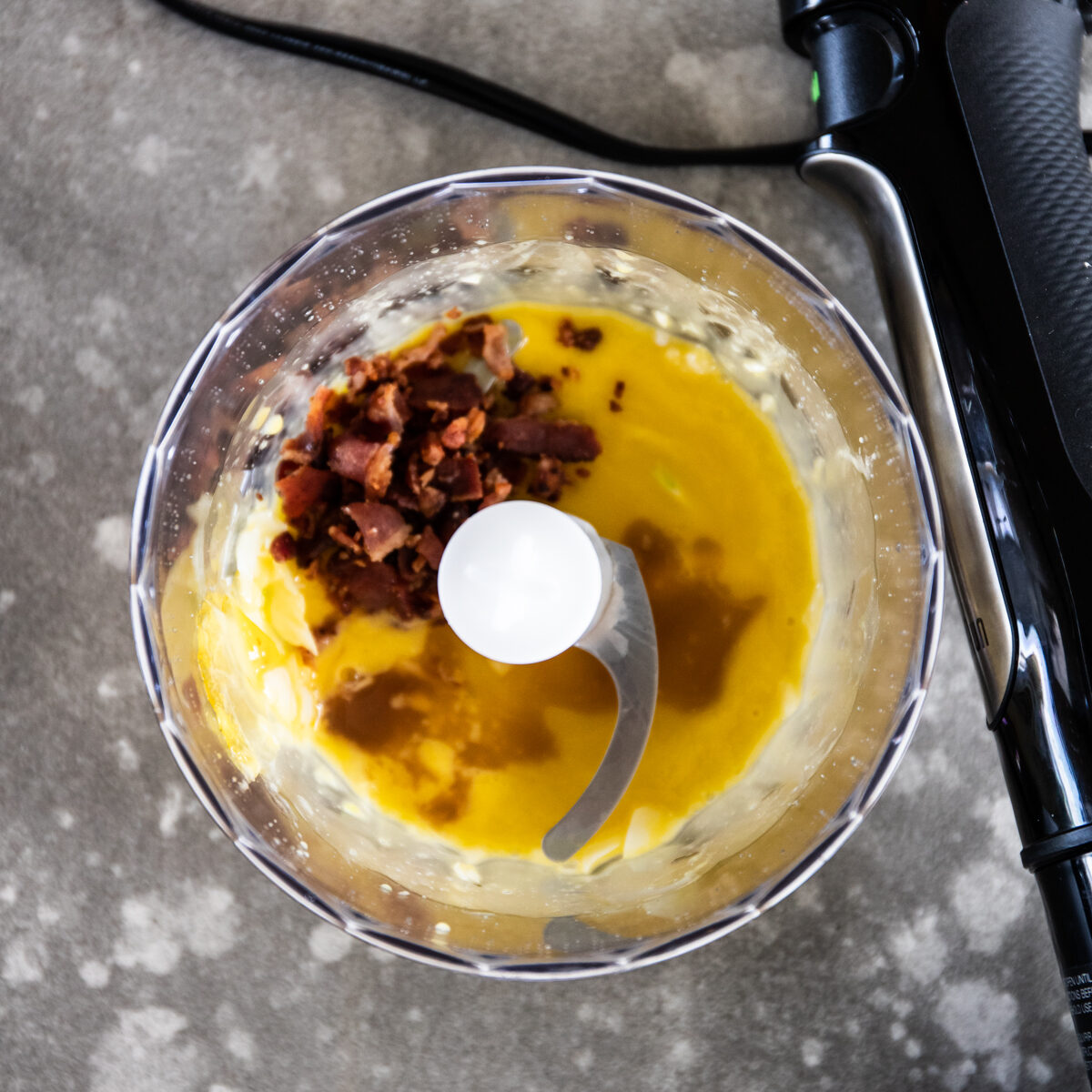 The yellow mixture has become frothy and bacon bits have been added on one side of the processor bowl.