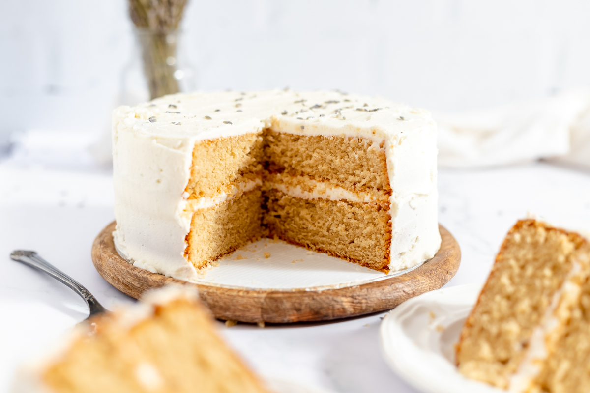 A round, tan colored 2 layer cake with creamy white frosting in between layers and on top. There is a wedge cut out of the front side of the cake and you can see the slices on plates in the foreground.