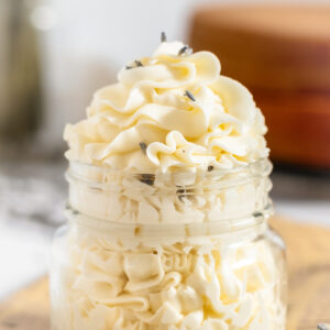 Closeup of creamy pale yellow white buttercream icing piped with a star tip into a clear glass jar with a few lavender buds sprinkled over.