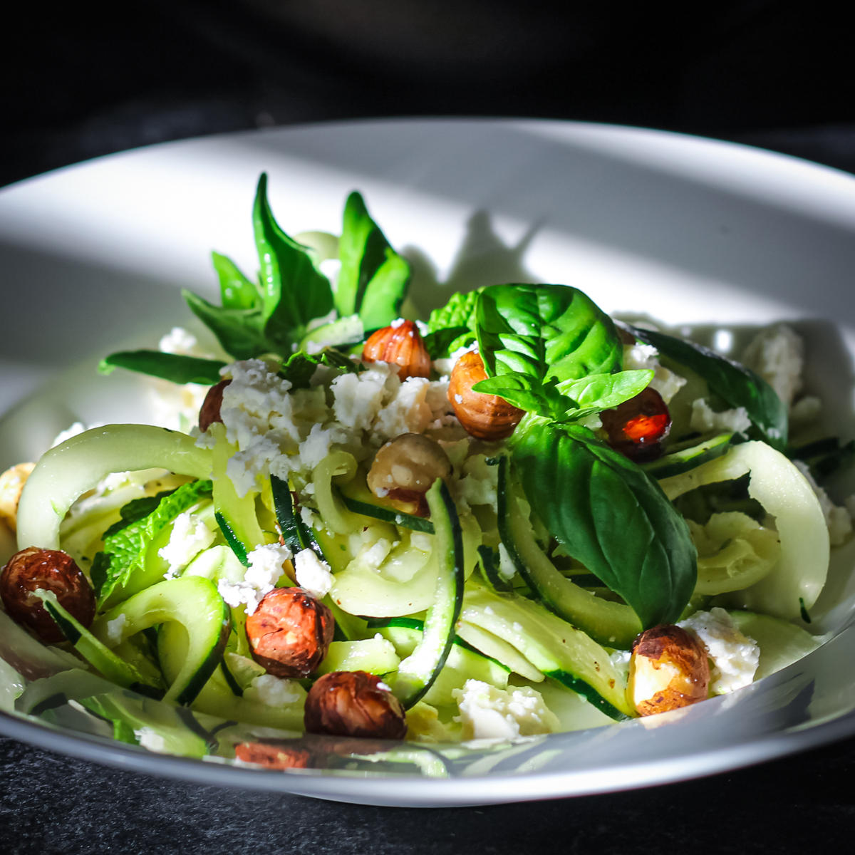 Raw zucchini feta salad with hazelnuts and basil leaves in a white bowl with shadow cast diagonally across.